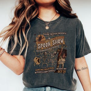 May include: Dark grey t-shirt with a vintage Halloween graphic. The design features the text "SPOOK SHOW" with illustrations of skeletons, a witch, and Frankenstein's monster. The shirt also includes the text "OCT 31".