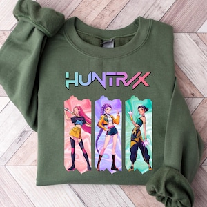 May include: Olive green sweatshirt with the word "HUNTRAX" in a colorful font. Below the text are three anime-style illustrations of women in action poses, each in a different outfit. The sweatshirt has a crew neck and long sleeves.