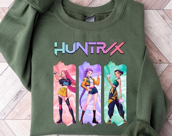 May include: Olive green sweatshirt with the word "HUNTRAX" in a colorful font. Below the text are three anime-style illustrations of women in action poses, each in a different outfit. The sweatshirt has a crew neck and long sleeves.