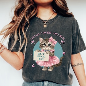 May include: Dark grey t-shirt with a cartoon kitten in a pink dress and bow, holding a sign saying "BUT FUCK ICE." The shirt also has the text "USUALLY SWEET AND NICE." The design is set against a blue circle.