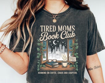Può includere: T-shirt grigio scuro con la scritta "TIRED MOMS Book Club" sopra un'illustrazione di un paesaggio urbano visto attraverso una finestra. La maglietta presenta anche la scritta "RUNNING ON COFFEE, CHAOS AND CHAPTERS".