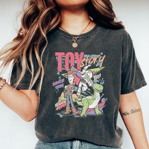 Puede incluir: Camiseta gris oscuro con un gr&aacute;fico colorido de los personajes de Toy Story, incluyendo a Woody, Buzz Lightyear y Rex. La camiseta tiene el texto "TOY STORY" y "SINCE 1995". La camiseta se combina con vaqueros azules y zapatos blancos.