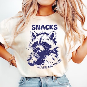 May include: A cream-coloured t-shirt featuring a navy blue raccoon illustration drinking from a cup. The words "SNACKS" and "MAKE ME NICER" are printed above and below the raccoon. The shirt is worn with light blue jeans.
