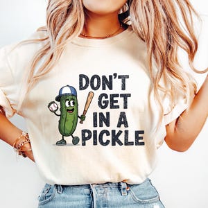May include: Cream-colored t-shirt featuring the phrase "DON'T GET IN A PICKLE" in black, with a cartoon pickle character holding a baseball bat and ball. The pickle wears a baseball cap. The shirt is paired with light blue jeans.