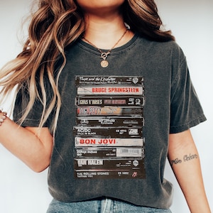 May include: Dark grey t-shirt with a print of stacked cassette tapes, including bands like Bruce Springsteen and The Rolling Stones. The person is wearing blue jeans and white trainers.