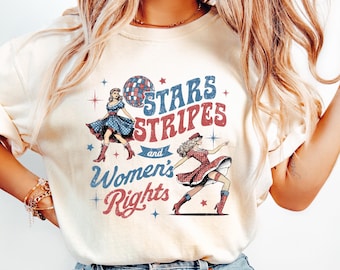 May include: Cream-colored t-shirt with the text "Stars Stripes and Women's Rights" in red and blue. The graphic features two dancing women in vintage dresses and a disco ball. The shirt is paired with light-wash denim jeans.