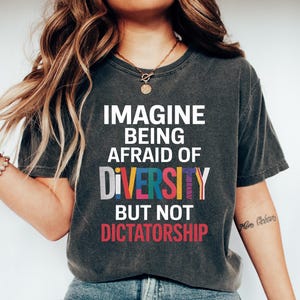 May include: Dark gray t-shirt with the text "IMAGINE BEING AFRAID OF DIVERSITY BUT NOT DICTATORSHIP" in white, red, yellow, blue, and green. The shirt is paired with blue jeans and white sneakers. The overall theme is social awareness.