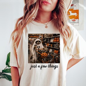 May include: A cream-colored short-sleeve t-shirt featuring a graphic of a raccoon dressed as a ghost, pushing a shopping cart filled with Halloween decorations in a store aisle. The text "just a few things" is printed in black below the graphic.