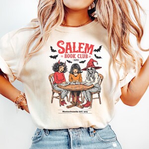 Puede incluir: Camiseta color crema con un gr&aacute;fico de tres figuras sentadas alrededor de una mesa, con el texto "Salem Book Club - 1692" y murci&eacute;lagos. Las figuras est&aacute;n vestidas como un diablo, una bruja y un esqueleto. Tambi&eacute;n est&aacute; presente el texto "Massachusetts Est. 1692".