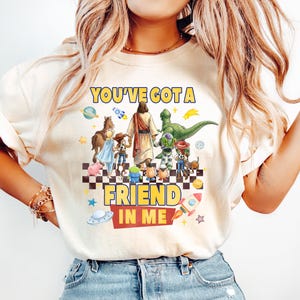 May include: A cream-colored short-sleeve t-shirt featuring a graphic print of a central figure walking with various cartoon characters on a checkered floor. The text reads YOU'VE GOT A FRIEND IN ME, surrounded by colorful space-themed icons and stars.