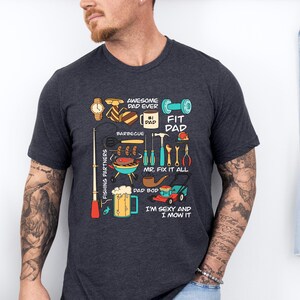 May include: Dark gray t-shirt with a colorful graphic design featuring tools, a barbecue, and the text "Awesome Dad Ever" and "Fit Dad". The shirt also includes the text "Mr. Fix It All" and "I'm Sexy and I Mow It".
