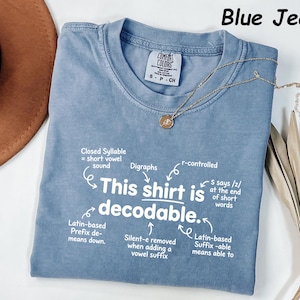 May include: A blue jean colored t-shirt with white text that reads This shirt is decodable. The graphic includes arrows pointing to linguistic notes such as Closed Syllable, Digraphs, r-controlled, and Latin-based prefixes and suffixes.