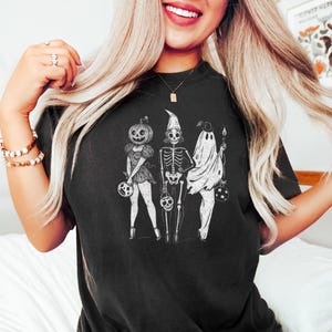 Może przedstawiać: Czarny t-shirt z grafiką o tematyce Halloween. Projekt przedstawia trzy postacie: postać z dyniową głową, szkielet i ducha, z kt&oacute;rych każdy trzyma wiadro na cukierki. Postacie są ustawione w rzędzie, w stylu vintage, lekko postarzanym.