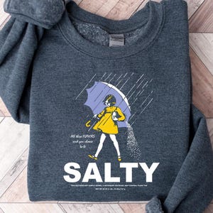 May include: Dark gray sweatshirt with a graphic of a person under a purple umbrella, wearing a yellow dress, and pouring salt. The word "SALTY" is printed in large white letters below the graphic. Text above the graphic reads "All the flavors and you choose to be."