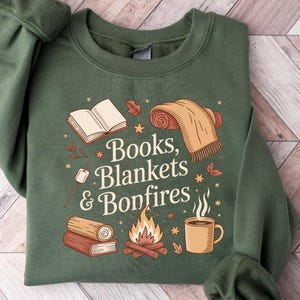 May include: Green sweatshirt with the text "Books, Blankets & Bonfires" and illustrations of a book, blanket, bonfire, and mug. The sweatshirt is a solid green color.