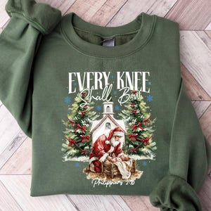 May include: An olive green sweatshirt featuring a Christmas-themed graphic. The design includes Santa Claus, a church, Christmas trees, and the text "EVERY KNEE Shall Bow" with "Philippians 2:10" below. The sweatshirt is laid flat on a wooden surface.