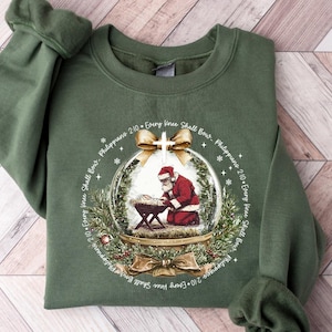 May include: Green sweatshirt featuring a snow globe design with Santa Claus kneeling before a manger. The snow globe is surrounded by festive decorations and the text "Every Knee Shall Bow - Philippians 2:10".