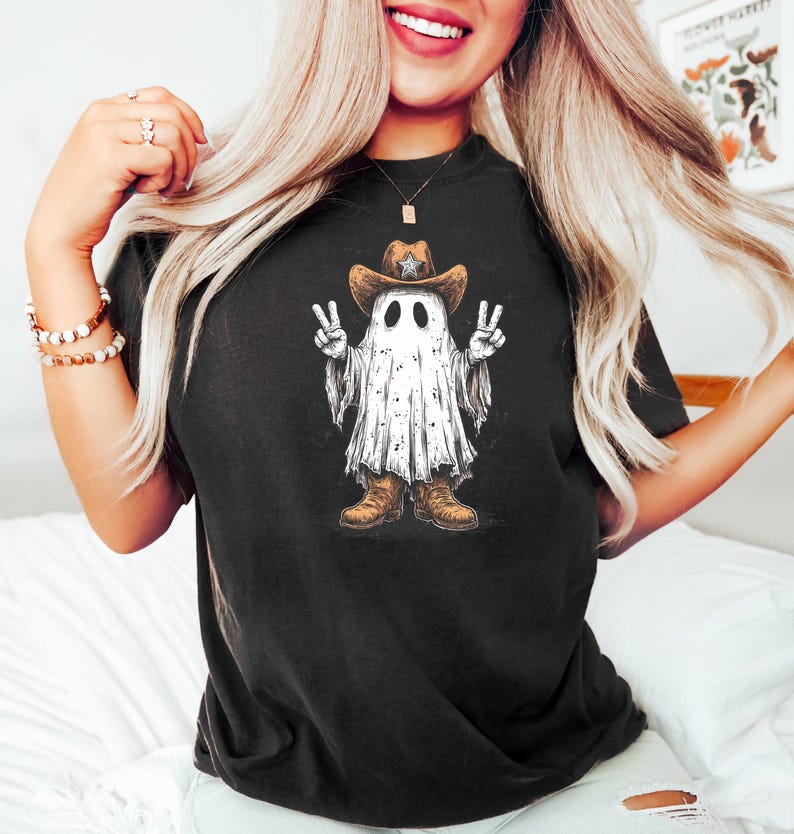 May include: Black t-shirt featuring a cartoon ghost wearing a cowboy hat and boots, making a peace sign with both hands. The ghost is white with black eyes and a distressed, textured design. The cowboy hat is brown with a star.