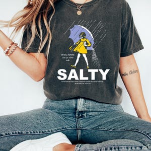 May include: Dark gray t-shirt with the word "SALTY" in white. The shirt features a graphic of a person in a yellow dress under a purple umbrella, with rain and salt pouring down. Text on the shirt reads "All the flavors and you choose to be."