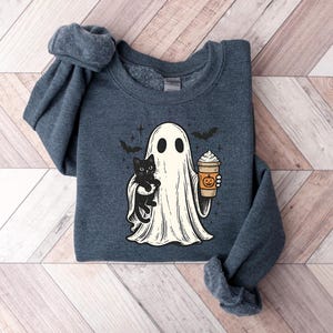 May include: A dark blue sweatshirt with a Halloween graphic. The design features a ghost holding a black cat and a pumpkin spice latte. Two bats and stars are also included. The sweatshirt is made of a soft material.