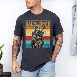 K&ouml;nnte beinhalten: Dunkelgraues T-Shirt mit einer Grafik der Figur The Mandalorian. Das Design enth&auml;lt den Text "THE GADALORIAN" &uuml;ber einem farbenfrohen gestreiften Hintergrund. Der Mandalorianer wird in voller R&uuml;stung dargestellt und h&auml;lt eine Waffe.