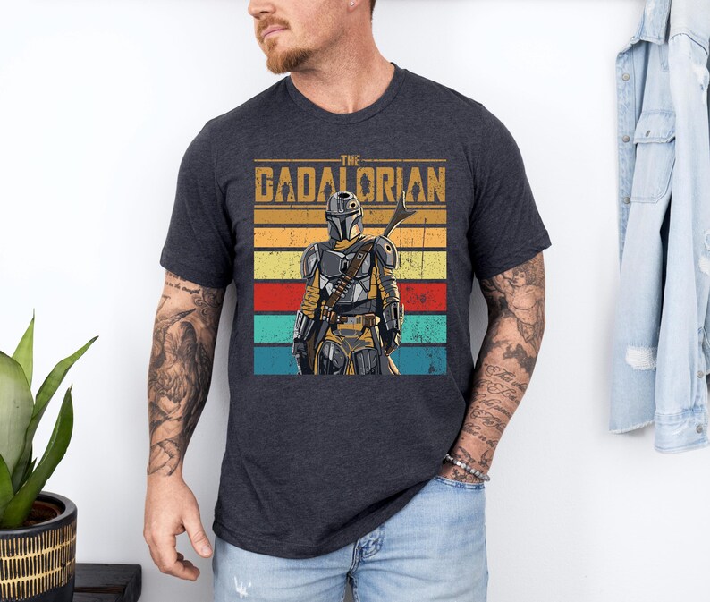 K&ouml;nnte beinhalten: Dunkelgraues T-Shirt mit einer Grafik der Figur The Mandalorian. Das Design enth&auml;lt den Text "THE GADALORIAN" &uuml;ber einem farbenfrohen gestreiften Hintergrund. Der Mandalorianer wird in voller R&uuml;stung dargestellt und h&auml;lt eine Waffe.