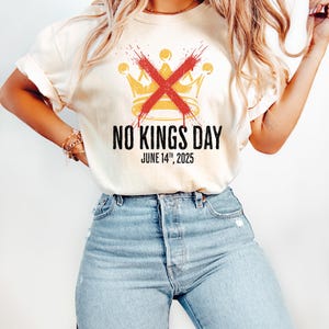 May include: Cream-colored t-shirt featuring a gold crown design crossed out with a red X. The text "NO KINGS DAY" and "JUNE 14TH, 2025" are printed below the crown in black. The shirt is worn with blue jeans.