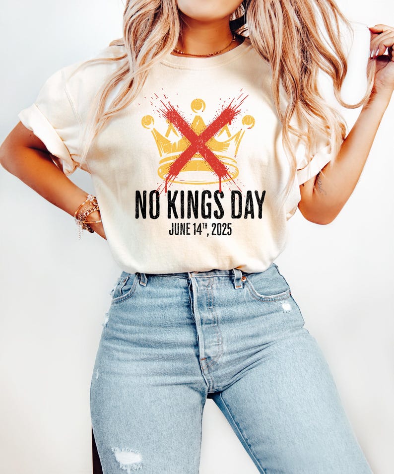 May include: Cream-colored t-shirt featuring a gold crown design crossed out with a red X. The text "NO KINGS DAY" and "JUNE 14TH, 2025" are printed below the crown in black. The shirt is worn with blue jeans.