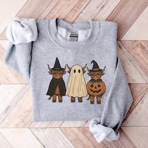 May include: A light gray sweatshirt featuring three cartoon Highland cows dressed for Halloween. One cow wears a witch hat and cape, another is a ghost, and the third is a pumpkin. The design is in shades of brown, black, and orange.