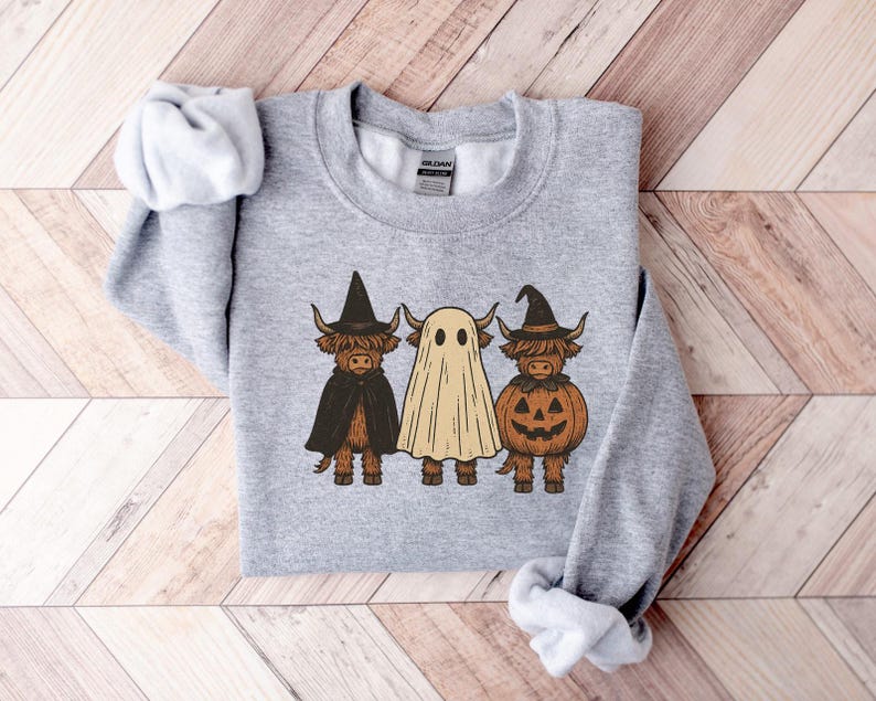 May include: A light gray sweatshirt featuring three cartoon Highland cows dressed for Halloween. One cow wears a witch hat and cape, another is a ghost, and the third is a pumpkin. The design is in shades of brown, black, and orange.