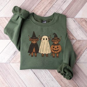May include: Green crewneck sweatshirt featuring a Halloween-themed graphic. The graphic depicts three Highland cows dressed as a witch, a ghost, and a jack-o'-lantern. The sweatshirt is made of a soft material and is perfect for the fall season.