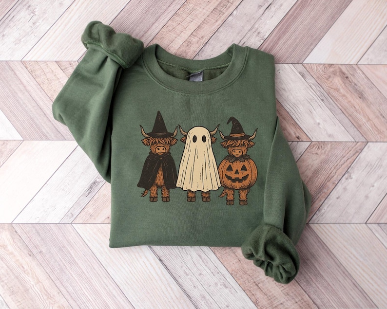 May include: Green crewneck sweatshirt featuring a Halloween-themed graphic. The graphic depicts three Highland cows dressed as a witch, a ghost, and a jack-o'-lantern. The sweatshirt is made of a soft material and is perfect for the fall season.