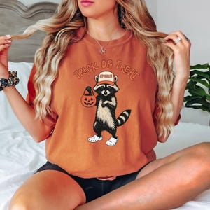 May include: Orange t-shirt featuring a raccoon wearing a hat and holding a jack-o'-lantern. The text "Trick or Treat" is arched above the raccoon. The raccoon is wearing white shoes. The hat says "Spooky".