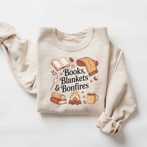 May include: Cream-colored sweatshirt with a graphic design featuring an open book, rolled blanket, campfire, and a mug. The text "Books, Blankets & Bonfires" is displayed in a decorative font. The sweatshirt has long sleeves.
