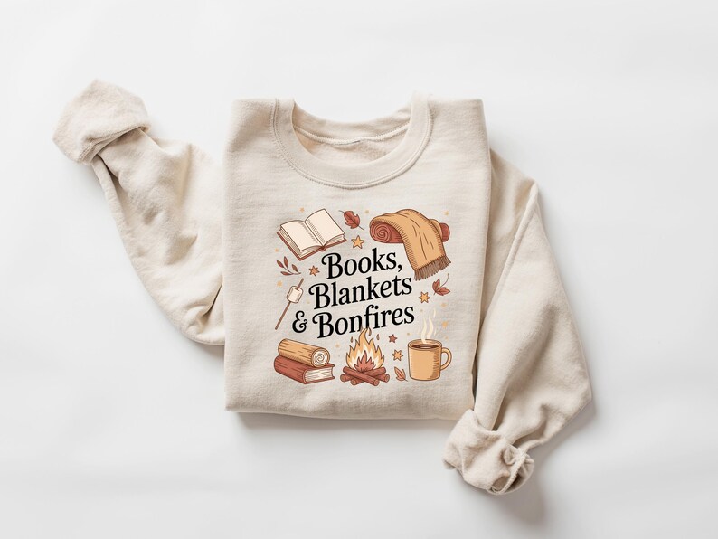 May include: Cream-colored sweatshirt with a graphic design featuring an open book, rolled blanket, campfire, and a mug. The text "Books, Blankets & Bonfires" is displayed in a decorative font. The sweatshirt has long sleeves.