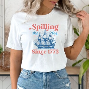 Może przedstawiać: Biała koszulka z grafiką niebieskiego i białego statku i tekstem "Spilling the Tea Since 1773".