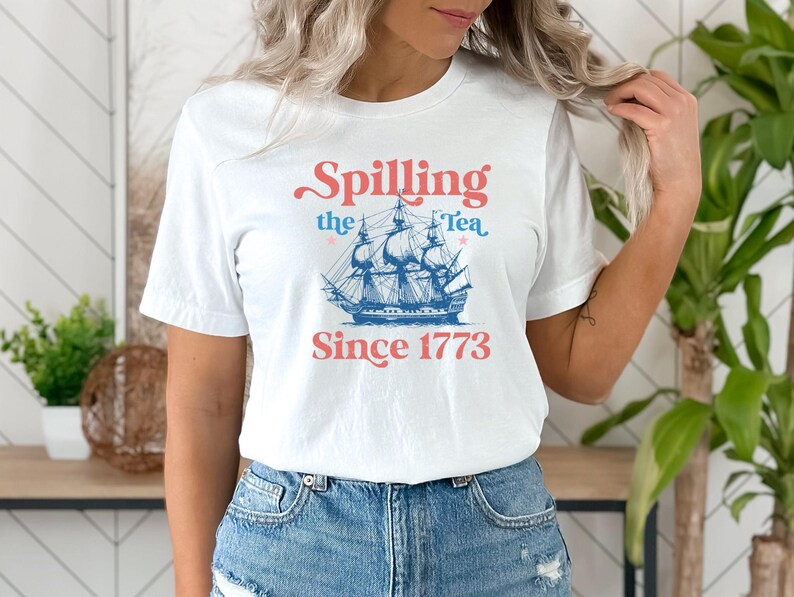 Może przedstawiać: Biała koszulka z grafiką niebieskiego i białego statku i tekstem "Spilling the Tea Since 1773".