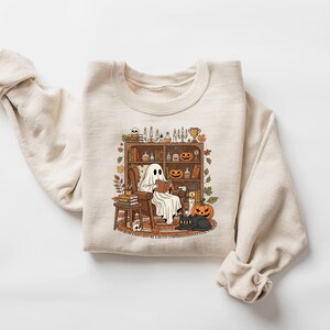 Peut inclure: Sweat-shirt cr&egrave;me avec un motif sur le th&egrave;me d'Halloween. Le dessin montre un fant&ocirc;me lisant dans un cadre confortable avec une biblioth&egrave;que, des citrouilles et un chat noir. Le sweat-shirt a des manches longues et un col rond.