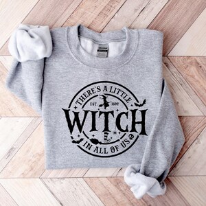 Op de afbeelding: Een lichtgrijs sweatshirt met de tekst "THERE'S A LITTLE WITCH IN ALL OF US" in een zwart cirkelvormig ontwerp. Het ontwerp bevat een heks op een bezem, vleermuizen en een heksenhoed. Het sweatshirt is op een houten oppervlak gevouwen.