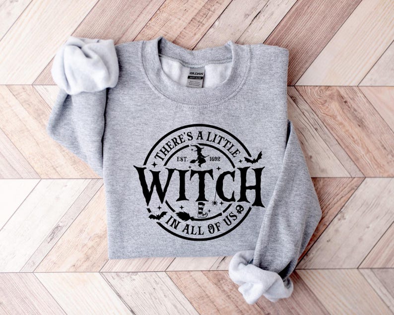 Op de afbeelding: Een lichtgrijs sweatshirt met de tekst "THERE'S A LITTLE WITCH IN ALL OF US" in een zwart cirkelvormig ontwerp. Het ontwerp bevat een heks op een bezem, vleermuizen en een heksenhoed. Het sweatshirt is op een houten oppervlak gevouwen.