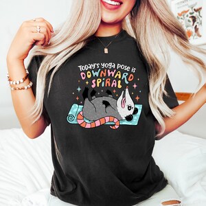 May include: Black t-shirt with a graphic of an opossum in a yoga pose on a blue mat. The text reads "Today's yoga pose is DOWNWARD SPIRAL" in colorful letters. The opossum is gray with a pink tail.