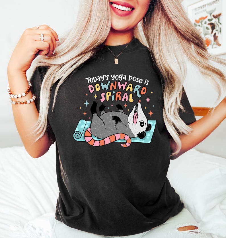 May include: Black t-shirt with a graphic of an opossum in a yoga pose on a blue mat. The text reads "Today's yoga pose is DOWNWARD SPIRAL" in colorful letters. The opossum is gray with a pink tail.