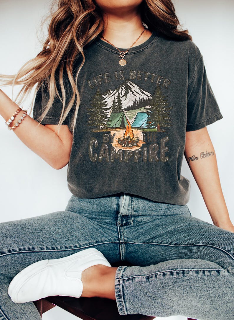 May include: A dark gray t-shirt with a graphic print of a mountain scene with a campfire, tents, and trees. The text "Life is better by the campfire" is printed on the shirt.