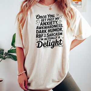 Pu&ograve; includere: Una maglietta color crema con testo nero che recita "Once You Get Past My Anxiety, Awkwardness, Dark Humor, RBF & Sarcasm I'm Actually A Delight".