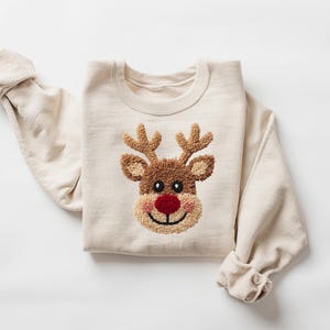 May include: A cream-colored sweatshirt featuring a reindeer face design. The reindeer has brown antlers, a brown face, a red nose, and a smiling mouth. The sleeves are rolled up.