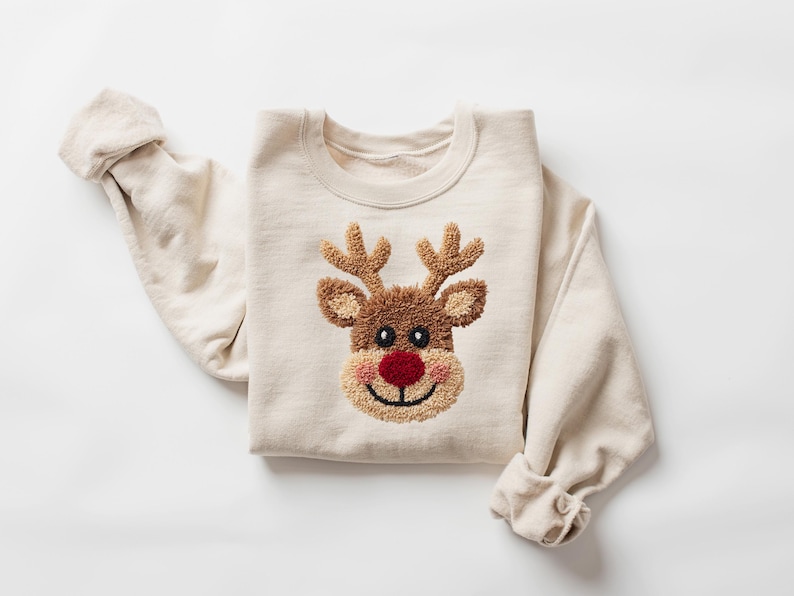 May include: A cream-colored sweatshirt featuring a reindeer face design. The reindeer has brown antlers, a brown face, a red nose, and a smiling mouth. The sleeves are rolled up.