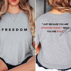 May include: A light gray t-shirt with the word "FREEDOM" printed in black on the front. The back of the shirt features the quote "JUST BECAUSE YOU ARE OFFENDED DOESN'T MEAN YOU ARE RIGHT" in black and red.