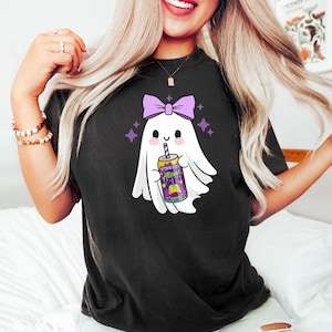 K&ouml;nnte beinhalten: Schwarzes T-Shirt mit einer Cartoon-Geisterfigur mit einer lila Schleife, die eine Dose Alan Energy Drink h&auml;lt. Der Geist hat ein l&auml;chelndes Gesicht und ist von lila Sternen umgeben. Die Dose ist lila und gelb.