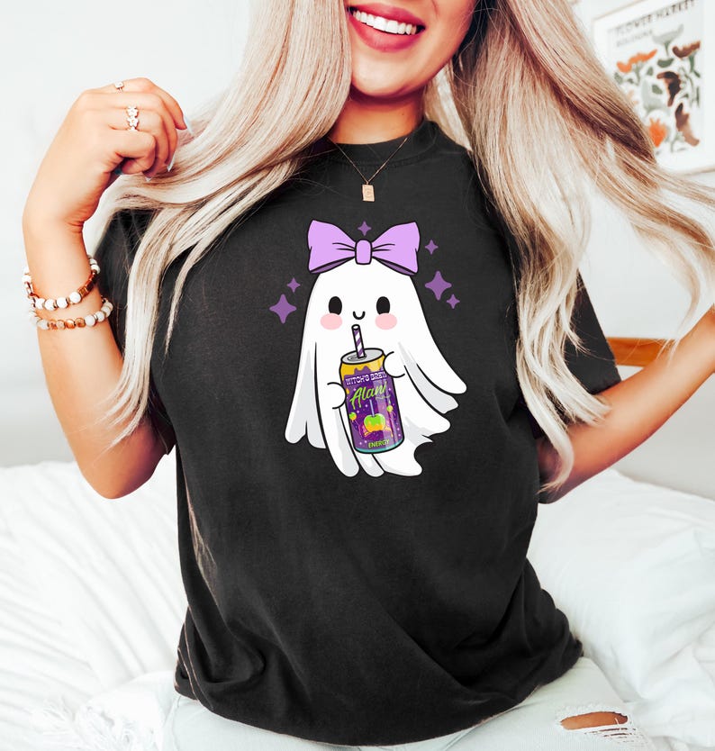 K&ouml;nnte beinhalten: Schwarzes T-Shirt mit einer Cartoon-Geisterfigur mit einer lila Schleife, die eine Dose Alan Energy Drink h&auml;lt. Der Geist hat ein l&auml;chelndes Gesicht und ist von lila Sternen umgeben. Die Dose ist lila und gelb.