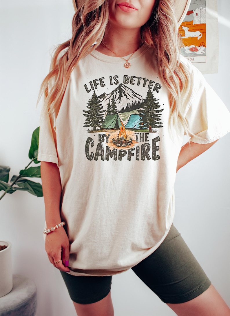 May include: A white t-shirt with a graphic print of a mountain scene with a campfire, tents, and trees. The text "Life is better by the campfire" is printed on the shirt.